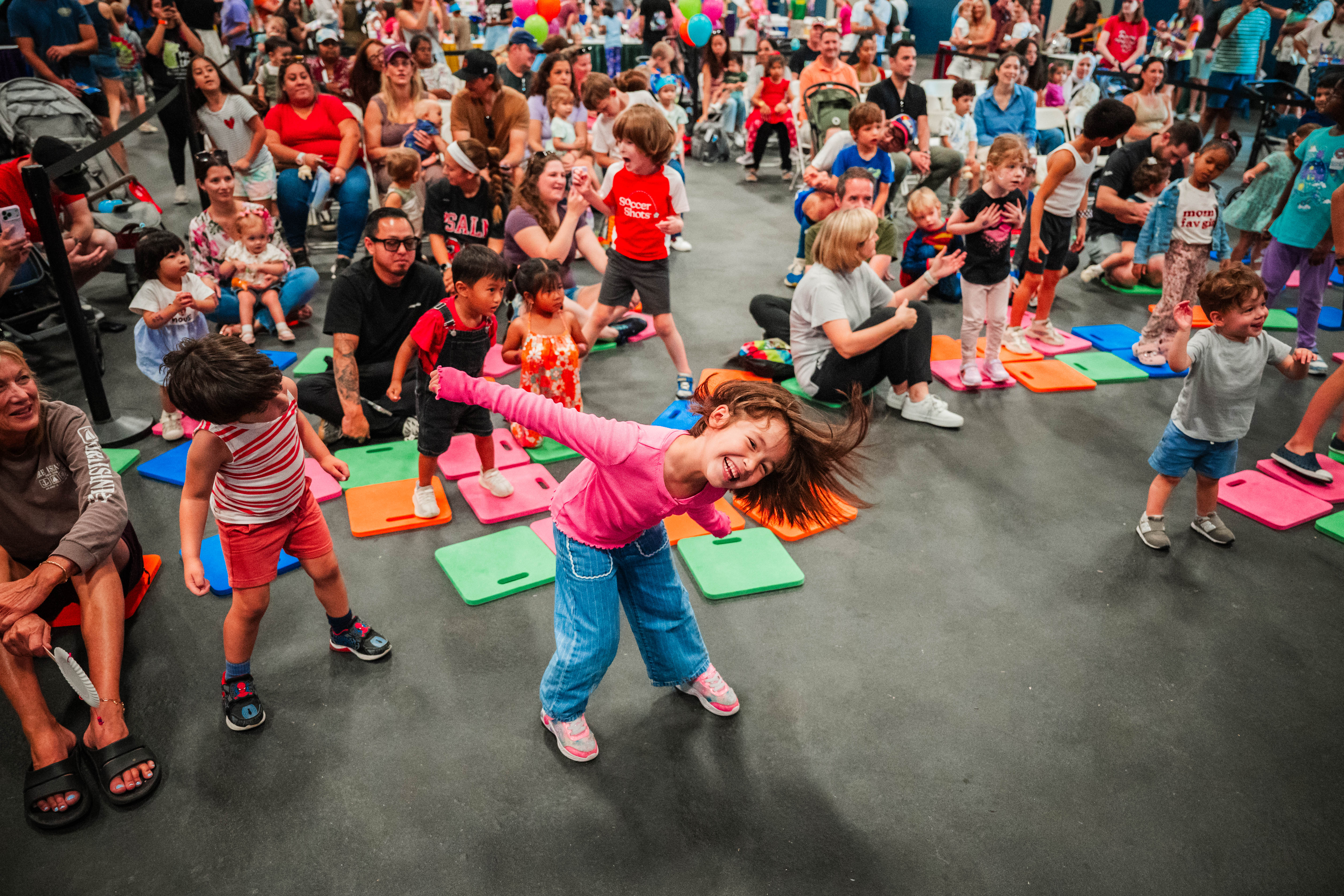 A young girl plays joyfully at the front of a large crowd near an indoor stage.