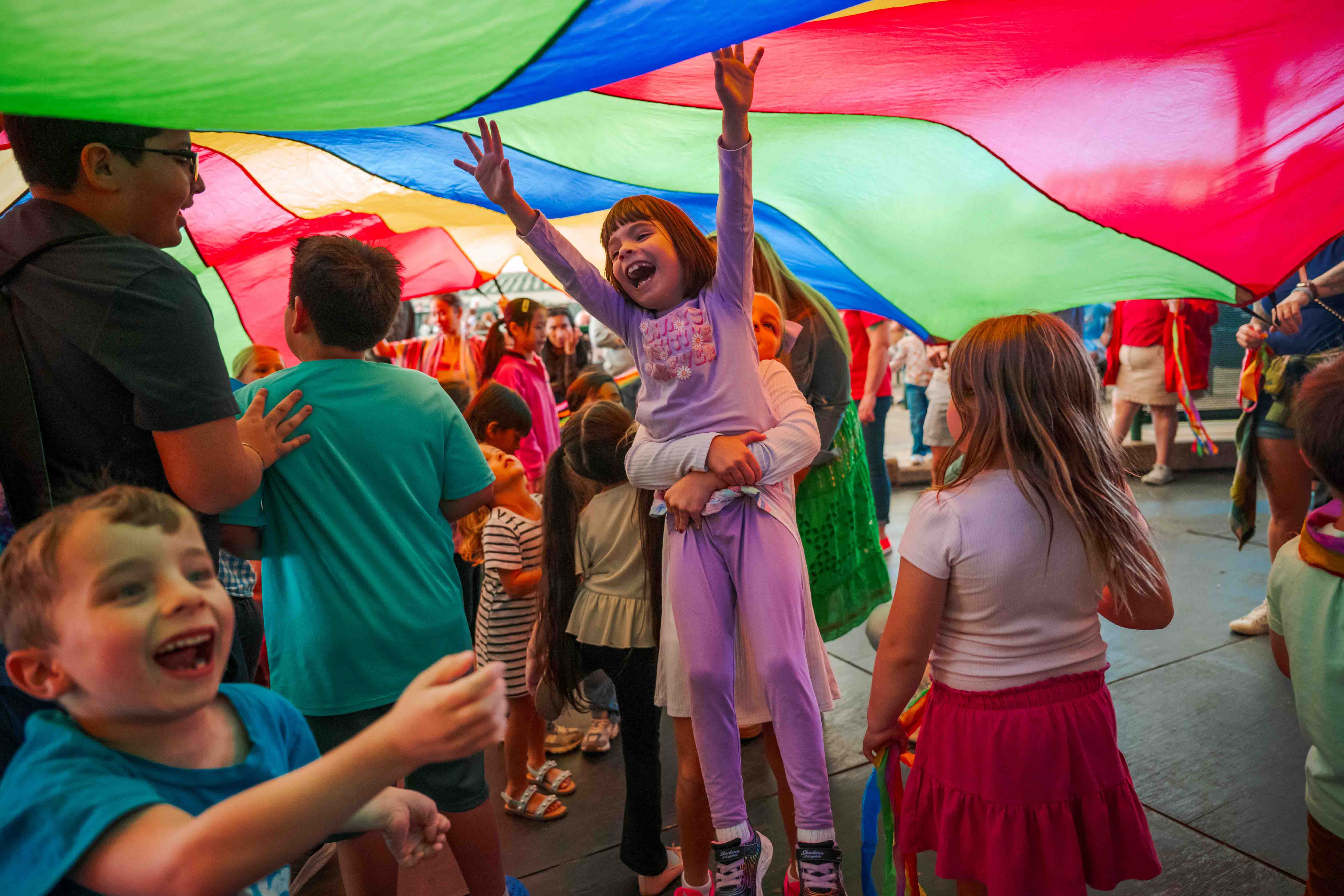 Children and families laugh together beneath a colorful play parachute.