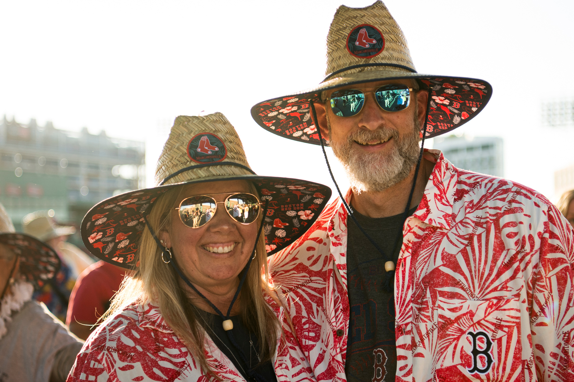A man and a woman smiling while wearing matching tropical shirts and straw hats with Boston Red Sox logos during a themed stadium event